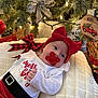 baby, holiday, christmas, red_bow, pacifier, pillow, christmas_tree, ornament, lights, decorations, festive, cute, indoor, smiling, child, infant, plaid_ribbon, wood_floor, cozy, dog