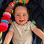 baby, smiling, blue_eyes, plush_toy, caterpillar_toy, stroller, clothing, beige_shirt, teal_pants, happy, child, cute, infant, person, indoor, portrait, playful, reaching_out, seat, pacifier