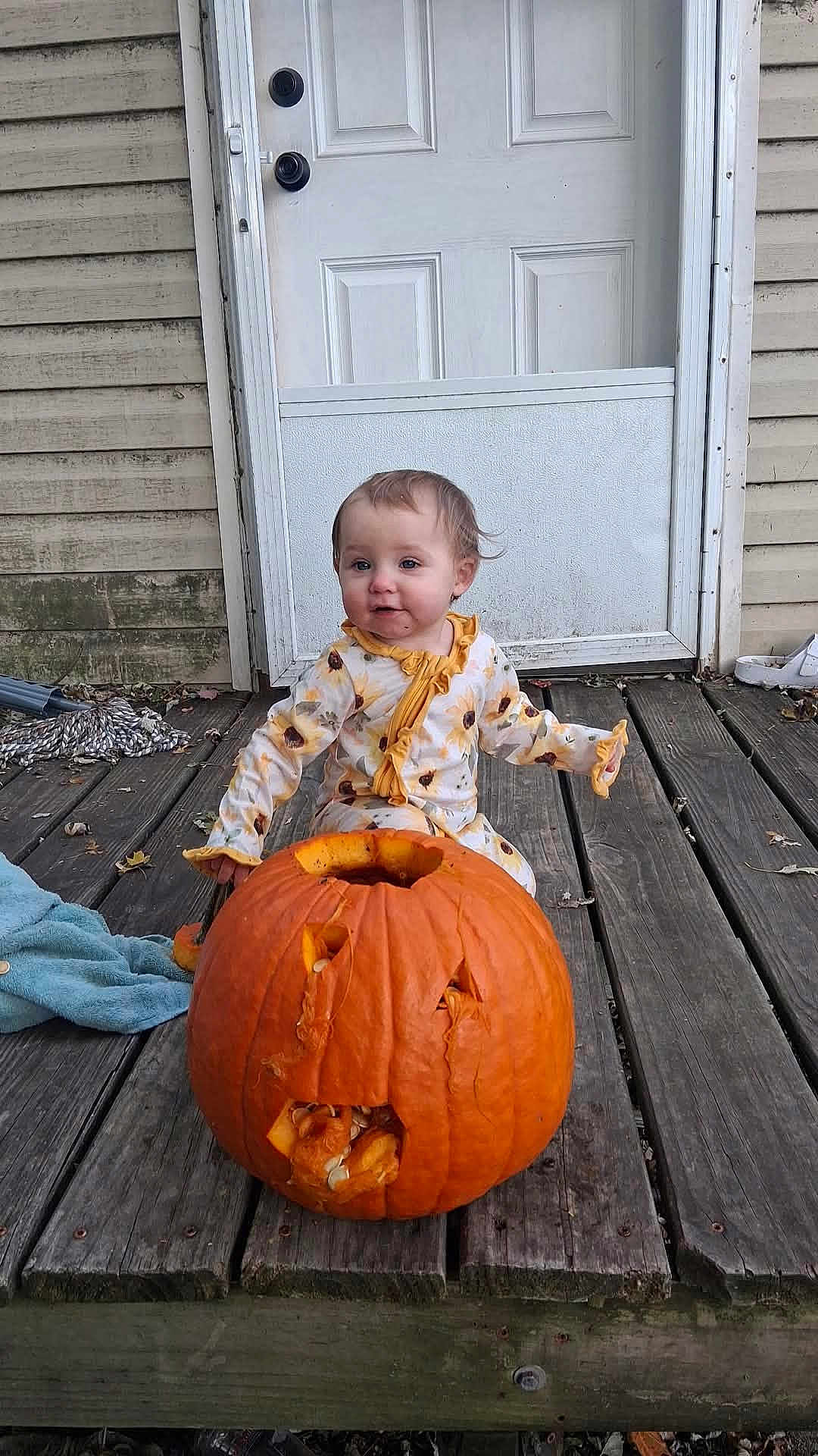 Blakeleigh Fisher is registered to the contest to win money with this photo: baby, child, toddler, pumpkin, jack_o_lantern, wooden_porch, door, pajamas, sunflower_pattern, fall, autumn, outdoor, person, face, smiling, curly_hair, door_handle, wood, seasonal, holiday