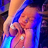 newborn, baby, sleeping, hospital, bassinet, infant, portrait, face, closed_eyes, hands, arm, medical_monitoring, electrodes, hospital_bracelet, blanket, blue_lighting, purple_lighting, peaceful, caregiver, indoor