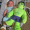 baby, infant, child, hulk, plush_toy, stuffed_animal, bouncer, baby_seat, sitting, smiling, portrait, face, indoor, carpet, shoes, romper, green, superhero, toy, nursery