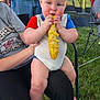 Charlie joined the competition — help win amazing prizes! baby, child, corn_on_the_cob, eating, outdoor, grass, person, sitting, lap, onesie, food, picnic, summer, casual, chair, adult, hand, footwear, tent, nature