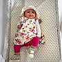 baby, infant, crib, floral_dress, hat, pink_leggings, boots, face, tongue_out, patterned_bedding, mesh_sides, cute, child, lying_down, playful, indoors, person, apparel, footwear, smiling