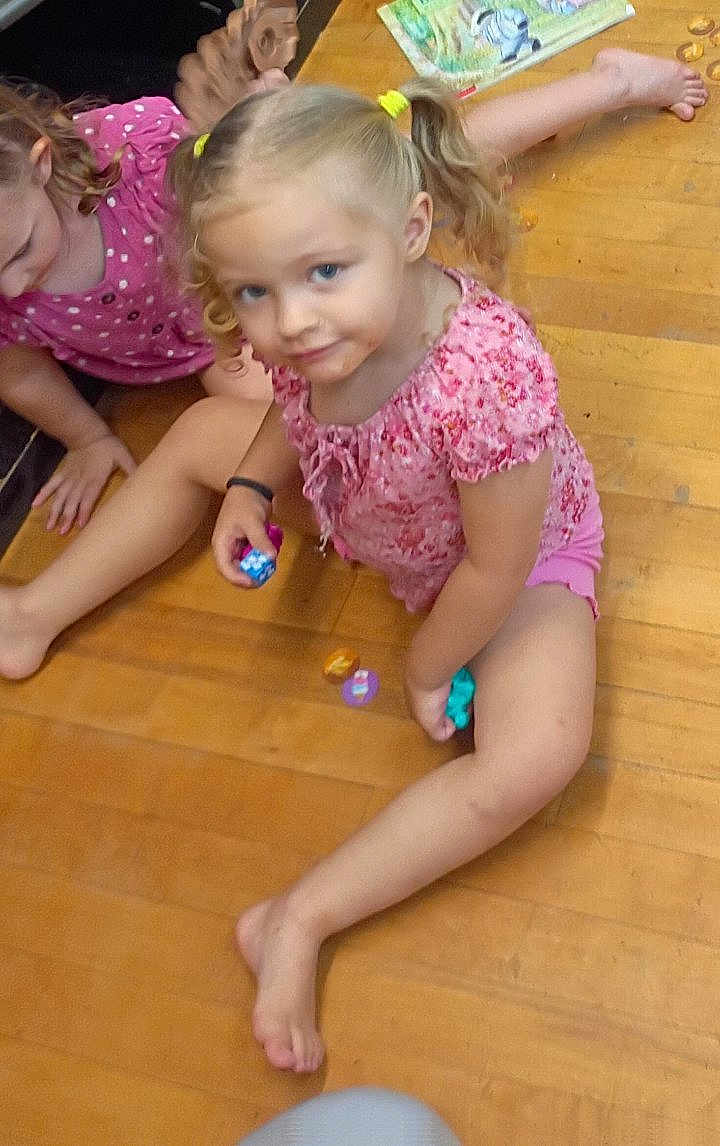 Jule is registered to the contest to win money with this photo: child, children, girl, floor, wooden_floor, toy, playing, indoor, casual, pink_clothing, pigtails, curly_hair, barefoot, book, colorful_toy, smile, sitting, person, young, playtime