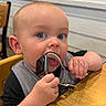 baby, child, blue_eyes, watch, smartwatch, chewing, table, high_chair, indoor, wooden_table, curious, face, hand, clothing, sitting, person, furniture, closeup, infant, cute