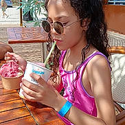 éloïse a rejoint le concours — aidez-le/la à gagner de superbes lots ! child, girl, sunglasses, ice_cream, cup, straw, pink_swimsuit, blue_wristband, wooden_table, outdoor, summer, beach_kiosk, curly_hair, chair, dessert, people, sunlight, vacation, portrait, eating