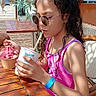 beach_kiosk, blue_wristband, chair, child, cup, curly_hair, dessert, eating, girl, ice_cream, outdoor, people, pink_swimsuit, portrait, straw, summer, sunglasses, sunlight, vacation, wooden_table