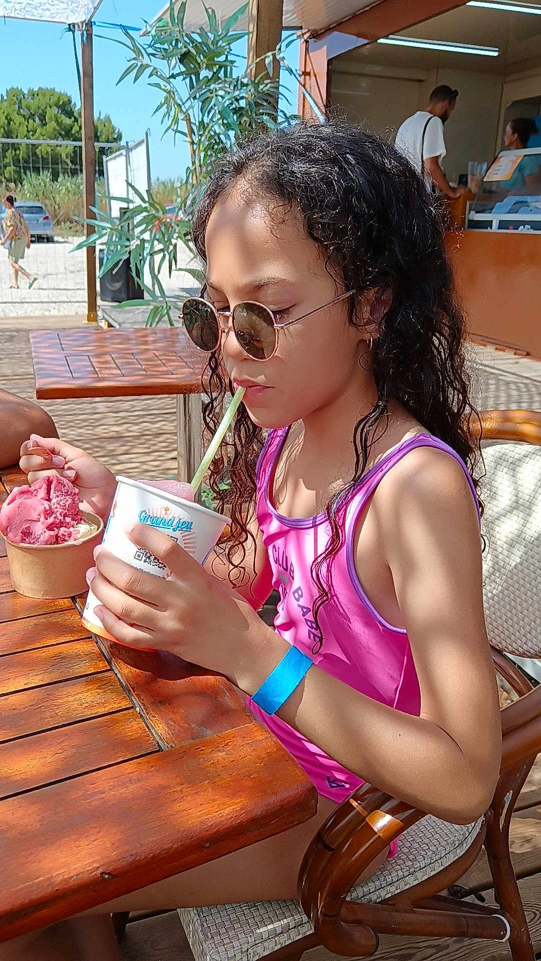 éloïse a rejoint le concours — aidez-le/la à gagner de superbes lots ! child, girl, sunglasses, ice_cream, cup, straw, pink_swimsuit, blue_wristband, wooden_table, outdoor, summer, beach_kiosk, curly_hair, chair, dessert, people, sunlight, vacation, portrait, eating