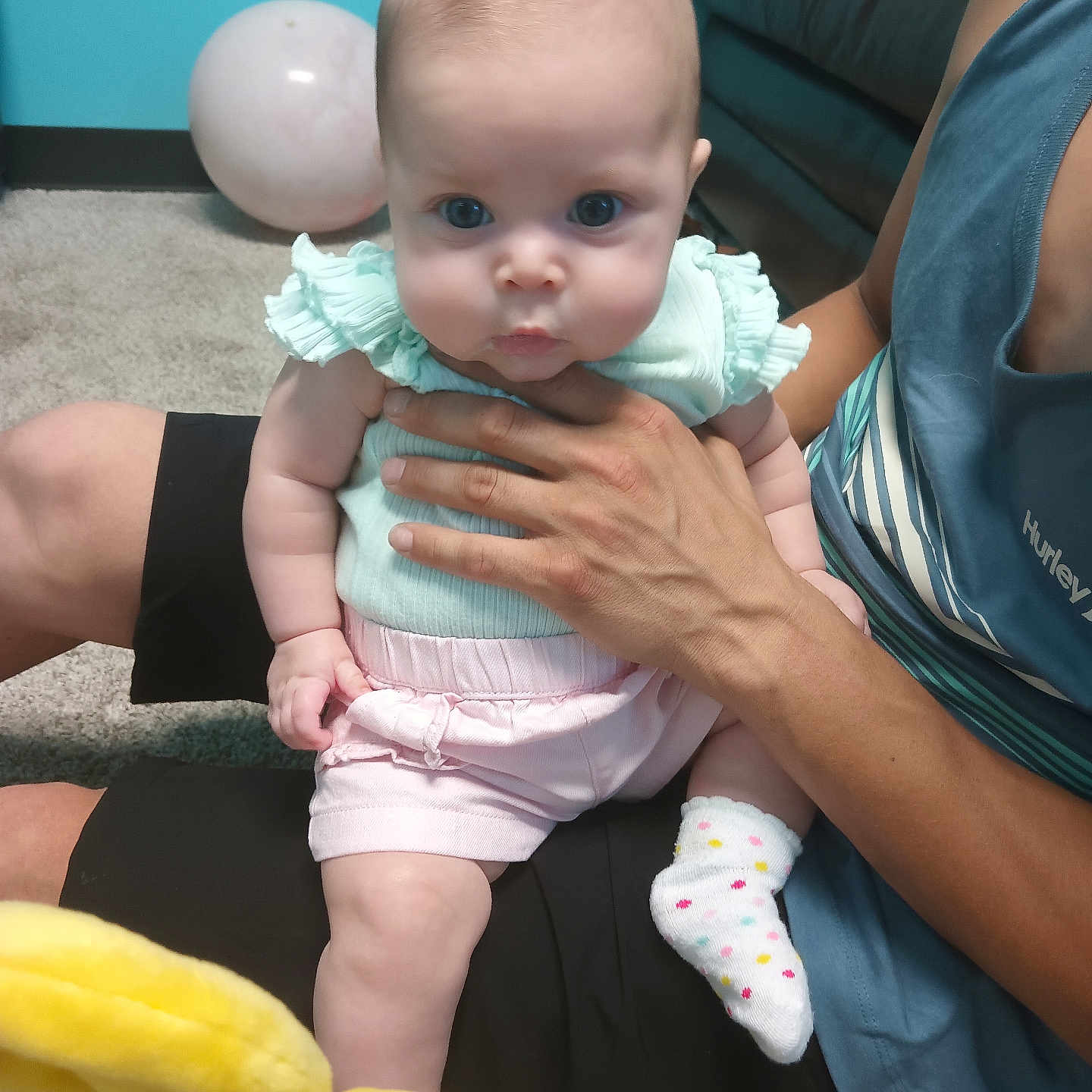 Poppie is registered to the contest to win money with this photo: adult, baby, balloon, blue_eyes, carpet, casual_clothing, child, couch, curious, hand, holding, indoor, mint_green, person, pink_shorts, plush_toy, polka_dot_socks, sitting, soft_toys, teal_wall