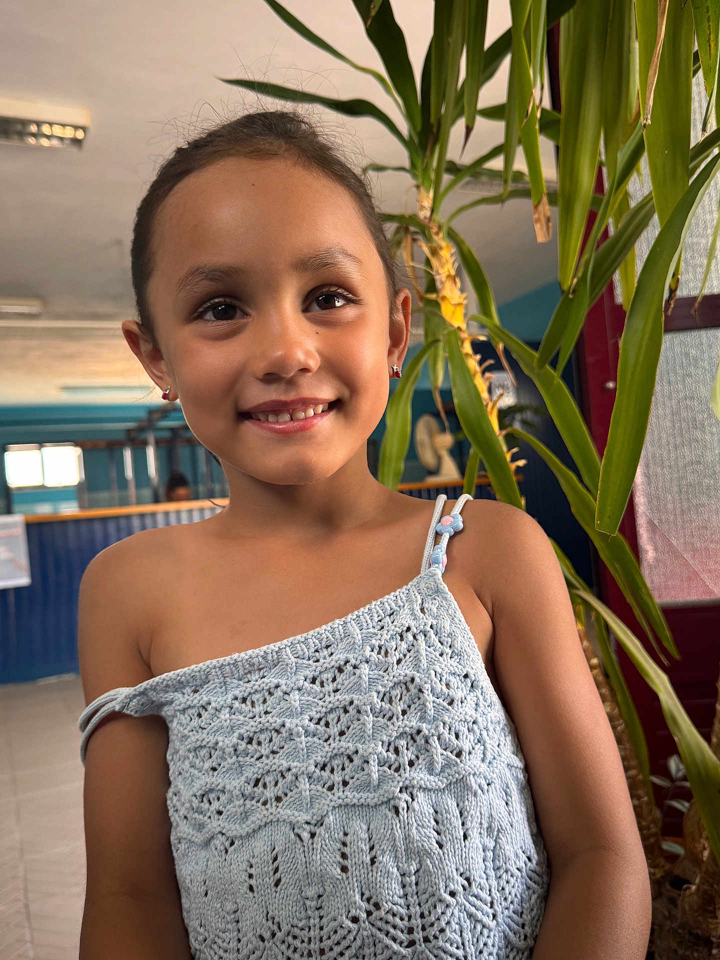 Hayden a rejoint le concours — aidez-le/la à gagner de superbes lots ! child, girl, smiling, portrait, indoor, knitted_top, earrings, plants, greenery, face, person, casual_clothing, happy, young, cute, hair_tied, background, light, beads, decor
