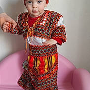 Tamila a rejoint le concours — aidez-le/la à gagner de superbes lots ! barefoot, bright_colors, child, colorful, curious, cute, fabric, hand_holding, hat, home, indoor, patterned_outfit, person, pink_chair, portrait, standing, textile, toddler, traditional_clothing, young_child