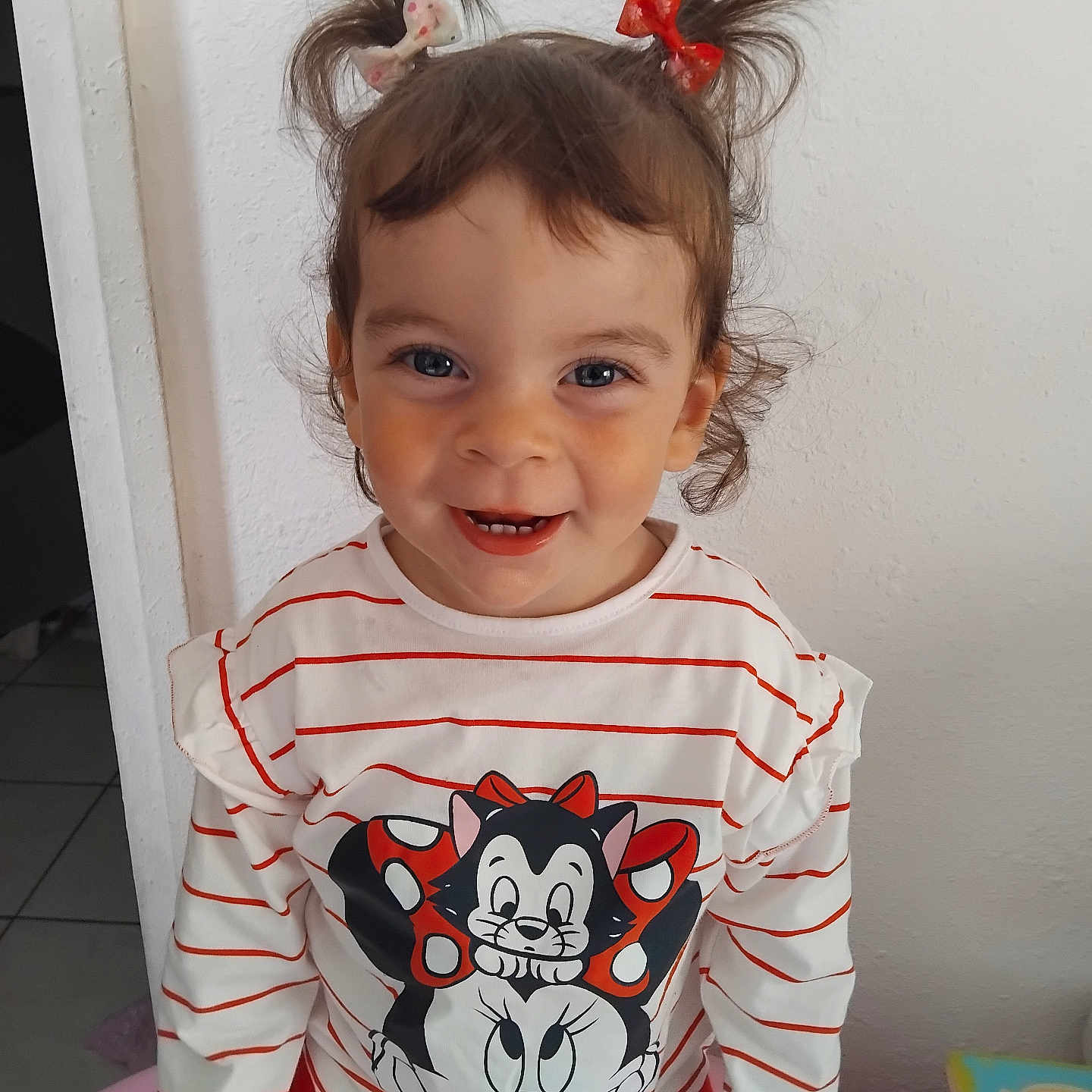 Tamila participe au concours pour gagner de l'argent avec cette photo : bright_colors, cartoon_character, casual_clothing, child, cute, face, hair_bows, happy, indoor, person, pigtails, playful, portrait, red_pants, smiling, standing, striped_shirt, toddler, white_wall, young_child