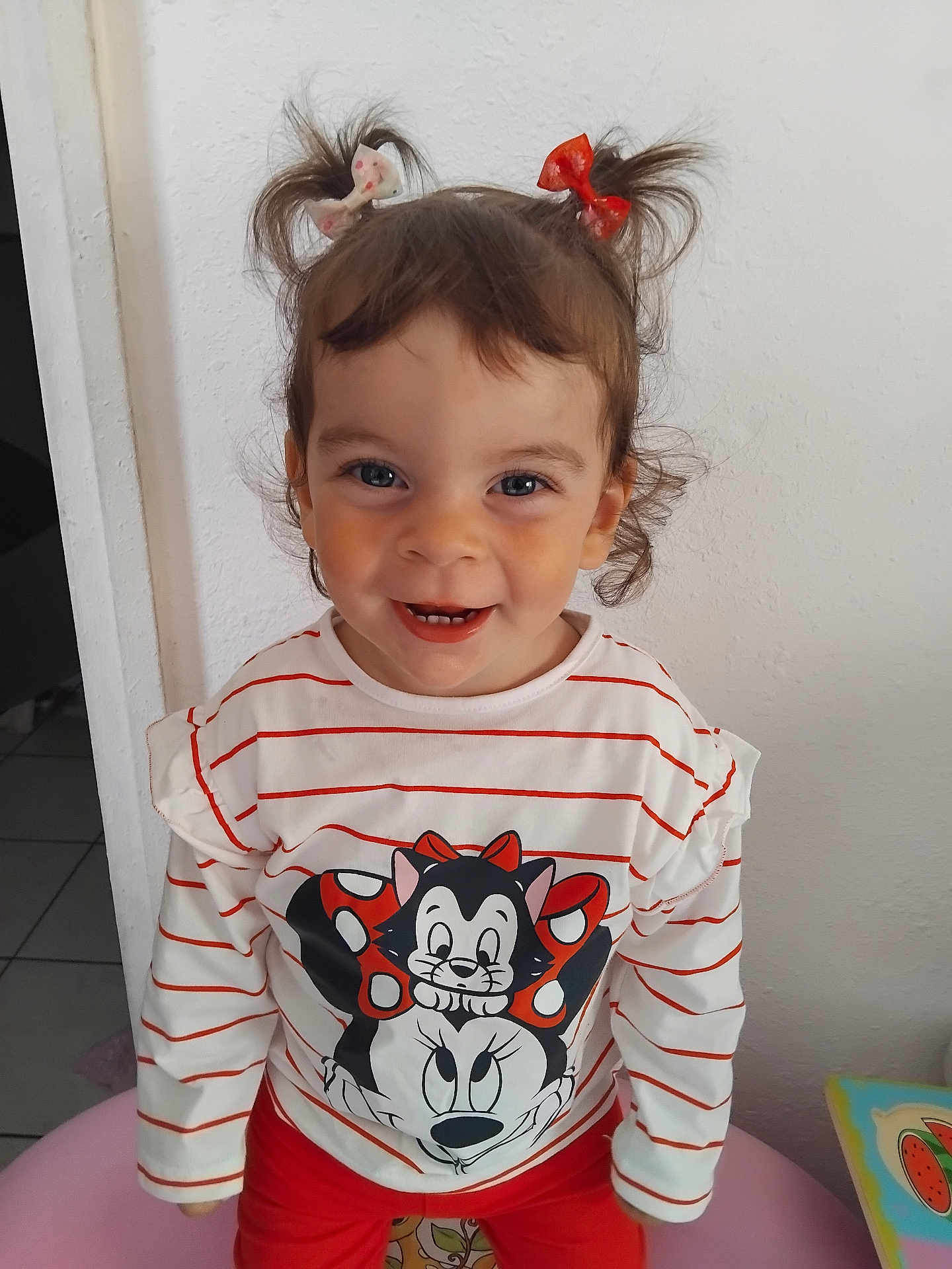 Tamila participe au concours pour gagner de l'argent avec cette photo : bright_colors, cartoon_character, casual_clothing, child, cute, face, hair_bows, happy, indoor, person, pigtails, playful, portrait, red_pants, smiling, standing, striped_shirt, toddler, white_wall, young_child