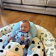 Aaron is registered to the contest to win money with this photo: baby, pajamas, plush_toy, baby_seat, cow_pattern, leaf_pattern, wooden_floor, couch, smile, infant, cute, indoor, comfort, child, person, blue_clothing, face, sitting, happy, soft_toy