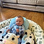 baby, pajamas, plush_toy, baby_seat, cow_pattern, leaf_pattern, wooden_floor, couch, smile, infant, cute, indoor, comfort, child, person, blue_clothing, face, sitting, happy, soft_toy