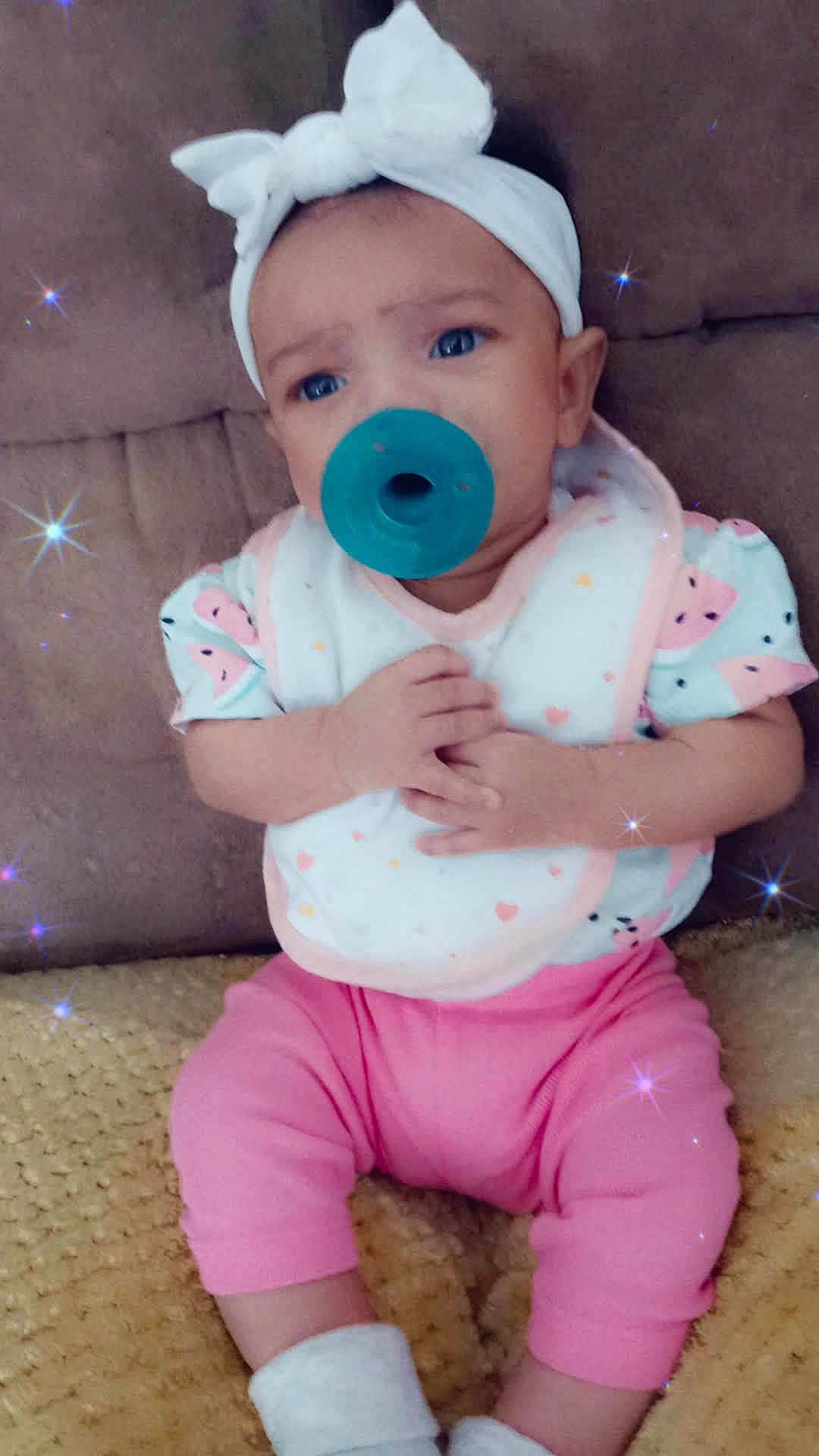 Tehlani Milele is registered to the contest to win money with this photo: baby, pacifier, headband, bow, pink_pants, white_socks, bib, couch, blanket, sparkle_effect, infant, sitting, cute, child, hands, face, clothing, indoor, portrait, soft_lighting