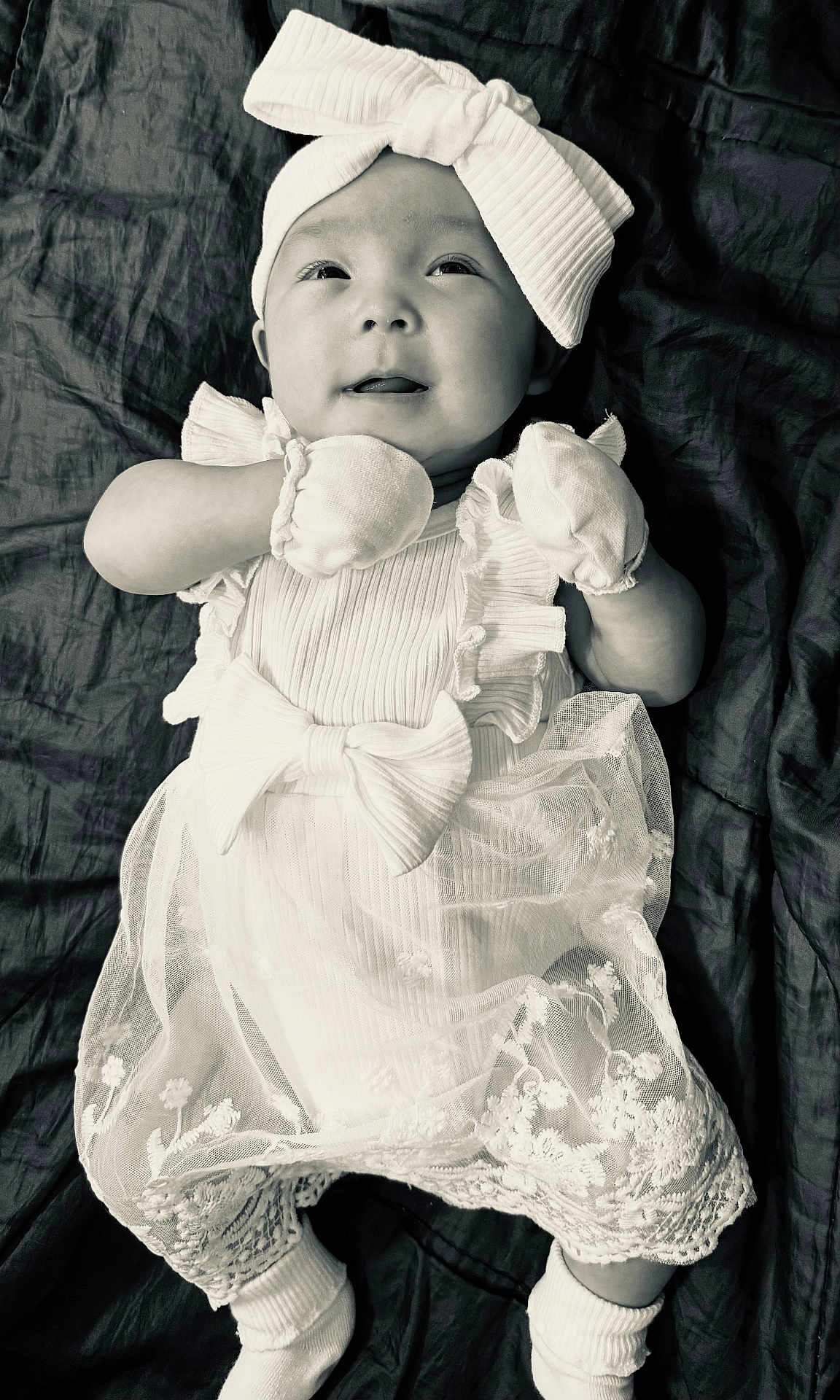 Emma joined the competition — help win amazing prizes! baby, infant, dress, bow, mittens, white_clothing, headband, portrait, laying_down, blanket, cute, child, smiling, soft_lighting, monochrome, fabric, young, expression, socks, newborn
