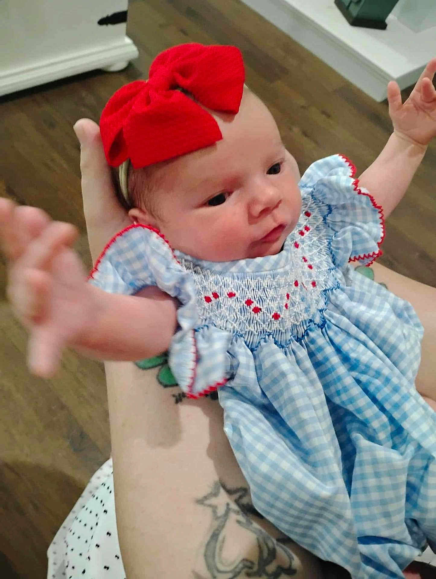 Jazmine is registered to the contest to win money with this photo: baby, infant, child, red_bow, blue_dress, tattoo, arm, hand, cute, indoor, wood_floor, person, face, smiling, holding, closeup, young, portrait, clothing, skin