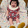 baby, smiling, infant, plaid_dress, yellow_shoes, white_collar, blanket, patterned_blanket, sitting, happy, cute, indoors, wooden_floor, child, person, young_child, black_hair, red_bow, sock, comfort