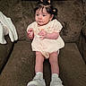 baby, child, sitting, couch, brown_couch, white_shoes, embroidered_dress, hair_buns, curly_hair, indoors, cute, infant, young_child, comfortable, clothing, portrait, person, legs, hands, soft_lighting