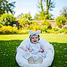 baby, child, smiling, dress, headband, bunny_ears, cushion, grass, flowers, garden, outdoor, sunny, greenery, happy, person, infant, nature, playful, cute, portrait