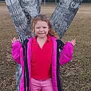 April is registered to the contest to win money with this photo: child, girl, tree, pink_jacket, pink_pants, smile, outdoor, grass, fall, nature, curly_hair, happy, playful, park, casual_clothing, young, person, posing, fun, seasonal
