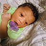baby, infant, child, portrait, lying_down, pacifier, green_outfit, striped_clothing, curly_hair, brown_eyes, smiling, awake, bed, blanket, bedspread, floral_pattern, indoor, closeup, hands, adorable