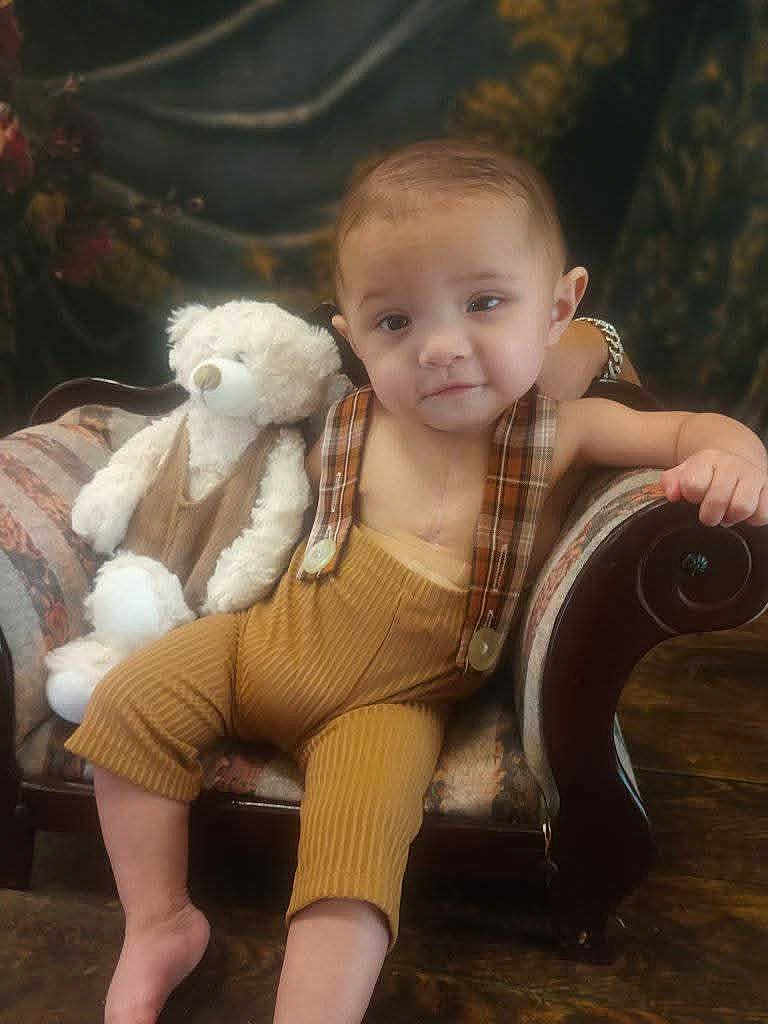 Timothy is registered to the contest to win money with this photo: child, toddler, teddy_bear, armchair, plaid, overalls, yellow, furniture, indoor, portrait, cute, baby, soft_toy, relaxed, smiling, floor, wooden_floor, background_blur, cozy, casual