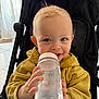 toddler, child, stroller, water_bottle, yellow_hoodie, blue_eyes, blond_hair, smiling, indoor, face, hand, clothing, cute, baby, person, seat, toy, playful, happy, portrait