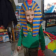 Maël a rejoint le concours — aidez-le/la à gagner de superbes lots ! board_games, bookshelf, cardboard_mask, child, coat, crafts, family_activity, green_hoodie, handmade_costume, happy, home, indoor, kid_fashion, living_room, pharaoh_headdress, playful, portrait, smile, television, toys