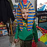 board_games, bookshelf, cardboard_mask, child, coat, crafts, family_activity, green_hoodie, handmade_costume, happy, home, indoor, kid_fashion, living_room, pharaoh_headdress, playful, portrait, smile, television, toys