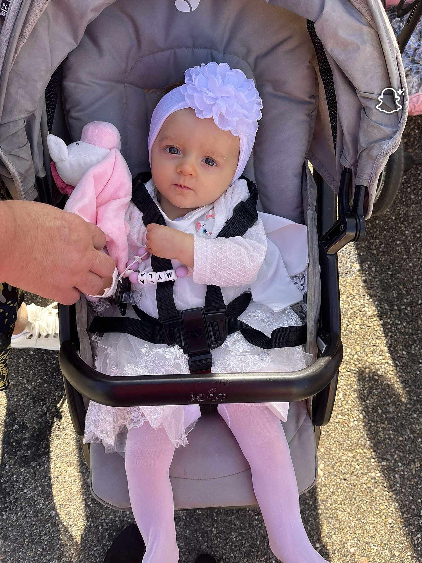 Myla a rejoint le concours — aidez-le/la à gagner de superbes lots ! baby, stroller, child, infant, white_dress, headband, flower, toy, hand, pink_tights, outdoor, sunlight, shadow, pavement, safety_harness, cute, portrait, person, seated, clothing