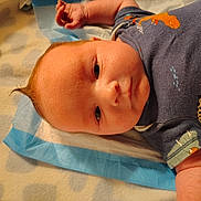Dylan is registered to the contest to win money with this photo: baby, infant, child, onesie, dinosaur_print, blanket, blue_pad, lying_down, closeup, face, skin, arm, hand, hair, expression, cute, newborn, indoors, soft_lighting, portrait
