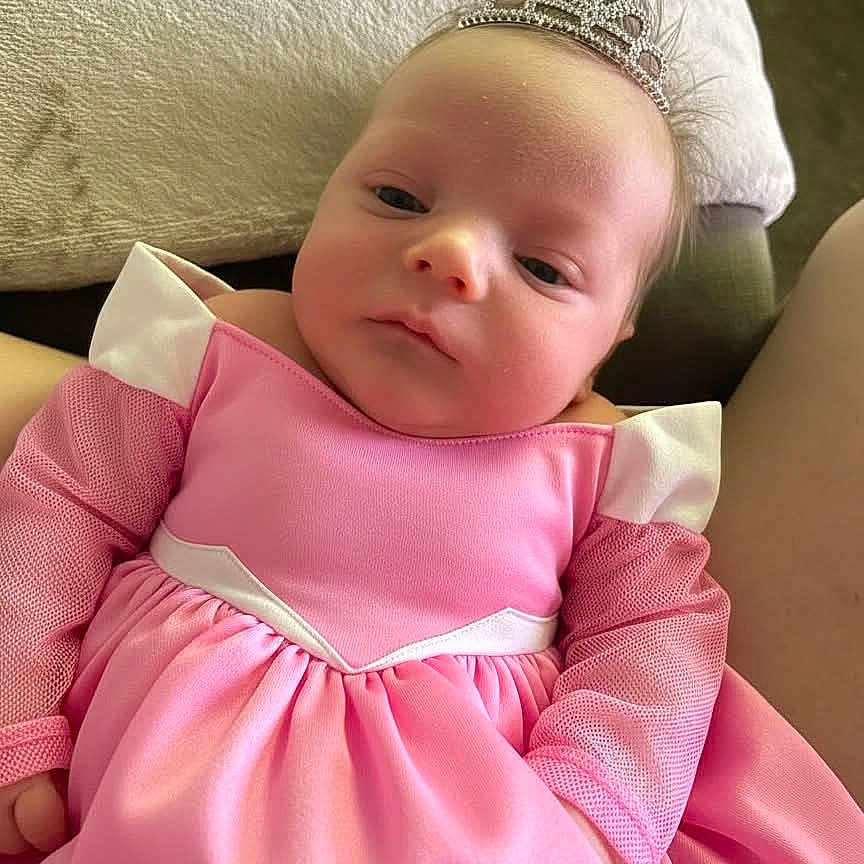 Ellie joined the competition — help win amazing prizes! baby, infant, tiara, pink_dress, mesh_sleeves, princess, cute, portrait, child, person, indoor, couch, blanket, clothing, headwear, young, small_hands, soft_light, relaxed, adorable