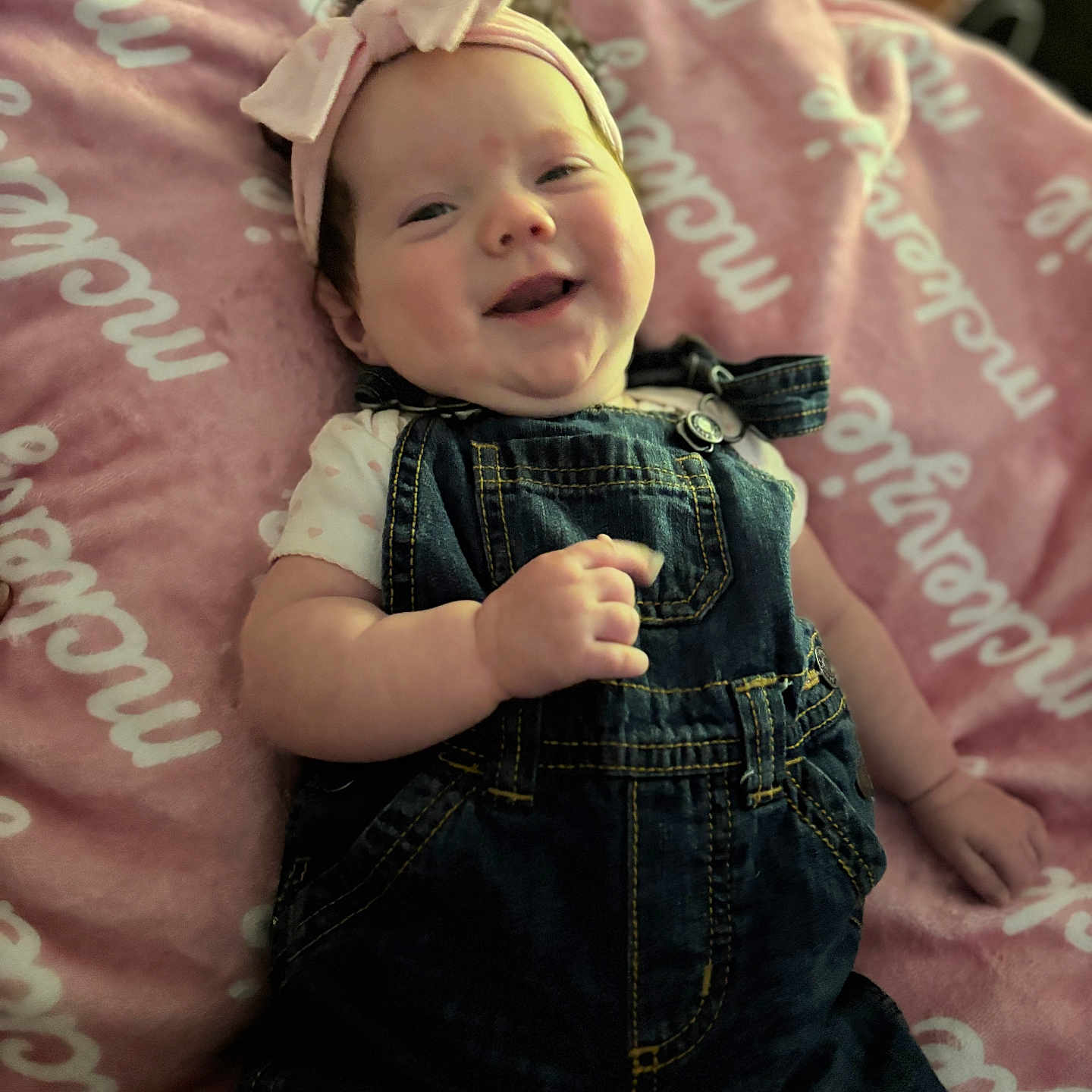 Mckenzie is registered to the contest to win money with this photo: baby, smiling, denim_overalls, headband, pink_bow, blanket, infant, happy, cute, child, portrait, clothing, soft_texture, lying_down, indoors, face, person, toddler, apparel, cozy