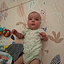 animal_pattern, baby, child, curious, expression, face, floor, hand, head, indoor, infant, legs, lying_down, onesie, pastel_colors, play_mat, small_toy, soft_lighting, striped_cloth, toy
