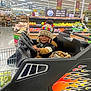 toddler, child, shopping_cart, grocery_store, hat, colorful, flames, black, produce, fruit, vegetables, smile, clothing, toy_car, indoor, aisle, shopping, person, cute, fun