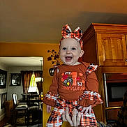 Aliyah joined the competition — help win amazing prizes! baby, child, smiling, indoor, thanksgiving, orange_clothing, plaid, headband, bow, kitchen, dining_room, furniture, hand, person, happy, portrait, holiday, celebration, infant, holding