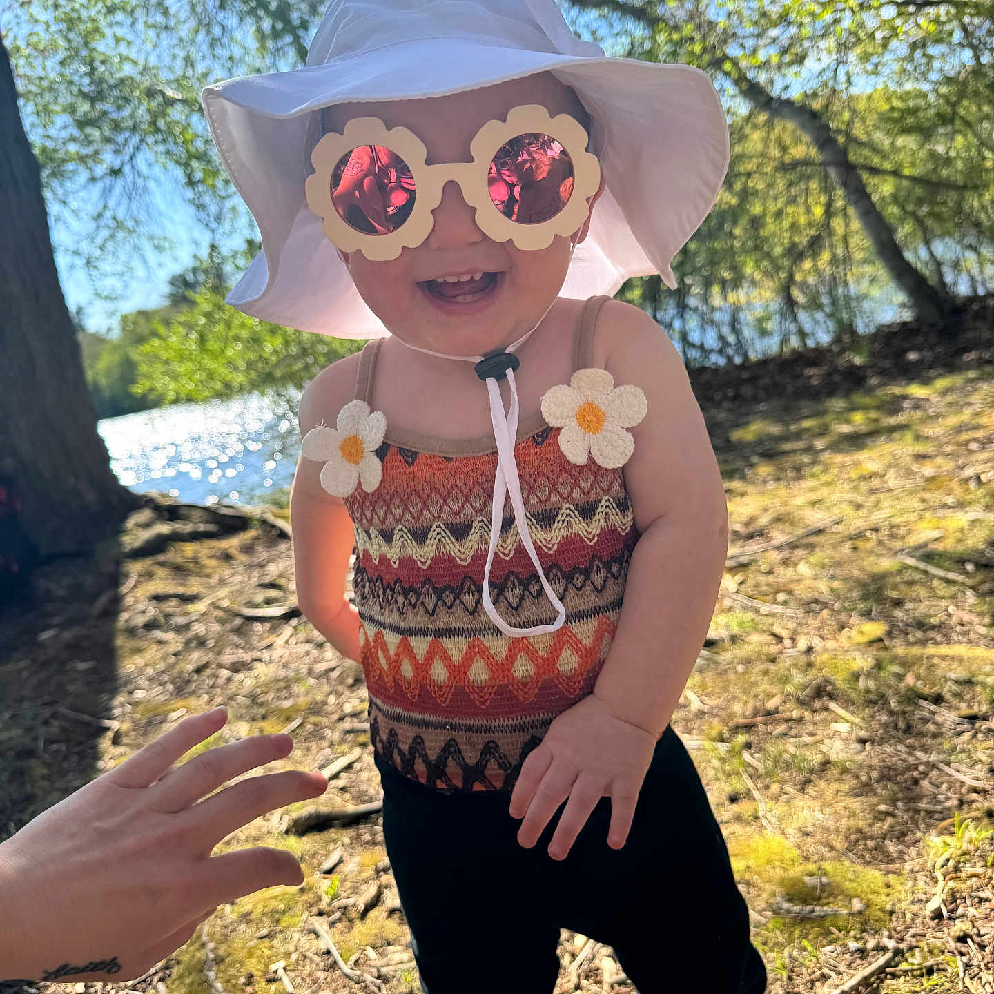 Aliyah is registered to the contest to win money with this photo: toddler, child, sunglasses, hat, outdoor, nature, sunlight, trees, lake, smile, happy, patterned_clothing, flower_accessory, playful, person, baby, summer, footwear, hand, shadow