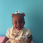 Tiana is registered to the contest to win money with this photo: baby, child, smiling, happy, sitting, girl, hair_accessories, bib, dress, patterned_skirt, indoor, portrait, blue_wall, cute, joyful, person, infant, clothing, hands, face