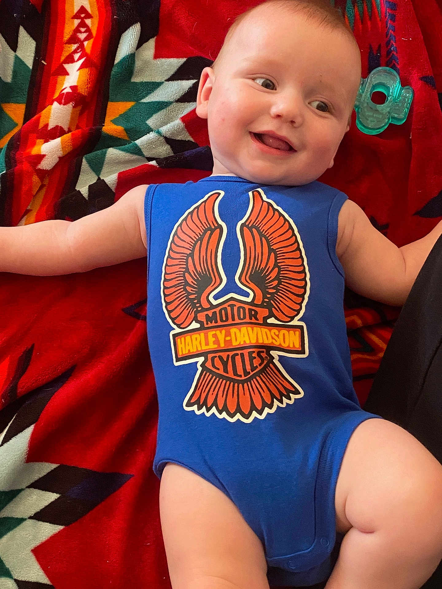 Carson joined the competition — help win amazing prizes! baby, infant, smile, blue_onesie, harley_davidson, blanket, patterned_blanket, teether, toy, lying_down, happy, face, arm, leg, skin, cute, child, portrait, indoor, clothing