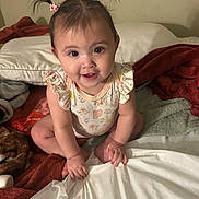 Xitlali is registered to the contest to win money with this photo: baby, child, pigtails, pink_hair_clips, bed, blankets, pillow, smile, happy, cute, indoor, person, young_child, sitting, hand, face, skin, clothing, infant, curious