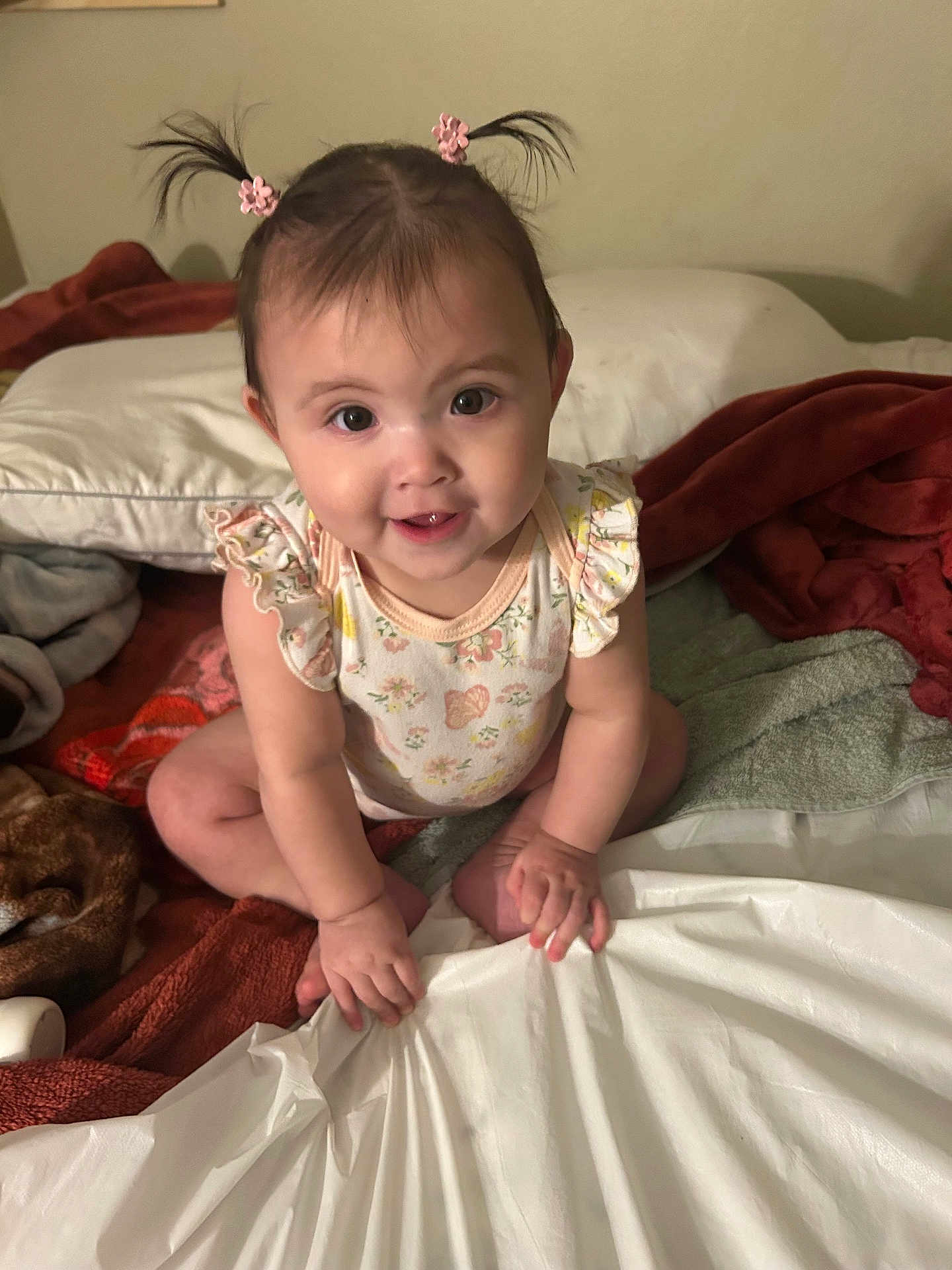 Xitlali is registered to the contest to win money with this photo: baby, child, pigtails, pink_hair_clips, bed, blankets, pillow, smile, happy, cute, indoor, person, young_child, sitting, hand, face, skin, clothing, infant, curious