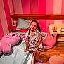 child, girl, bed, plush_toy, stuffed_animal, pajamas, braids, smile, pink_wall, wooden_floor, pillow, socks, slippers, furniture, room, toy, happy, indoor, person, colorful