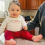 adult_arm, baby, bow, cabinet, child, countertop, foot, hair_accessory, happy, indoors, infant, kitchen, person, polka_dot, red_pants, sink, smiling, toaster, white_shirt, window