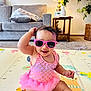 baby, child, pink_sunglasses, pink_tutu, play_mat, living_room, couch, table, lamp, plants, smiling, sitting, cute, indoors, toddler, fun, happy, toy, colorful, decor