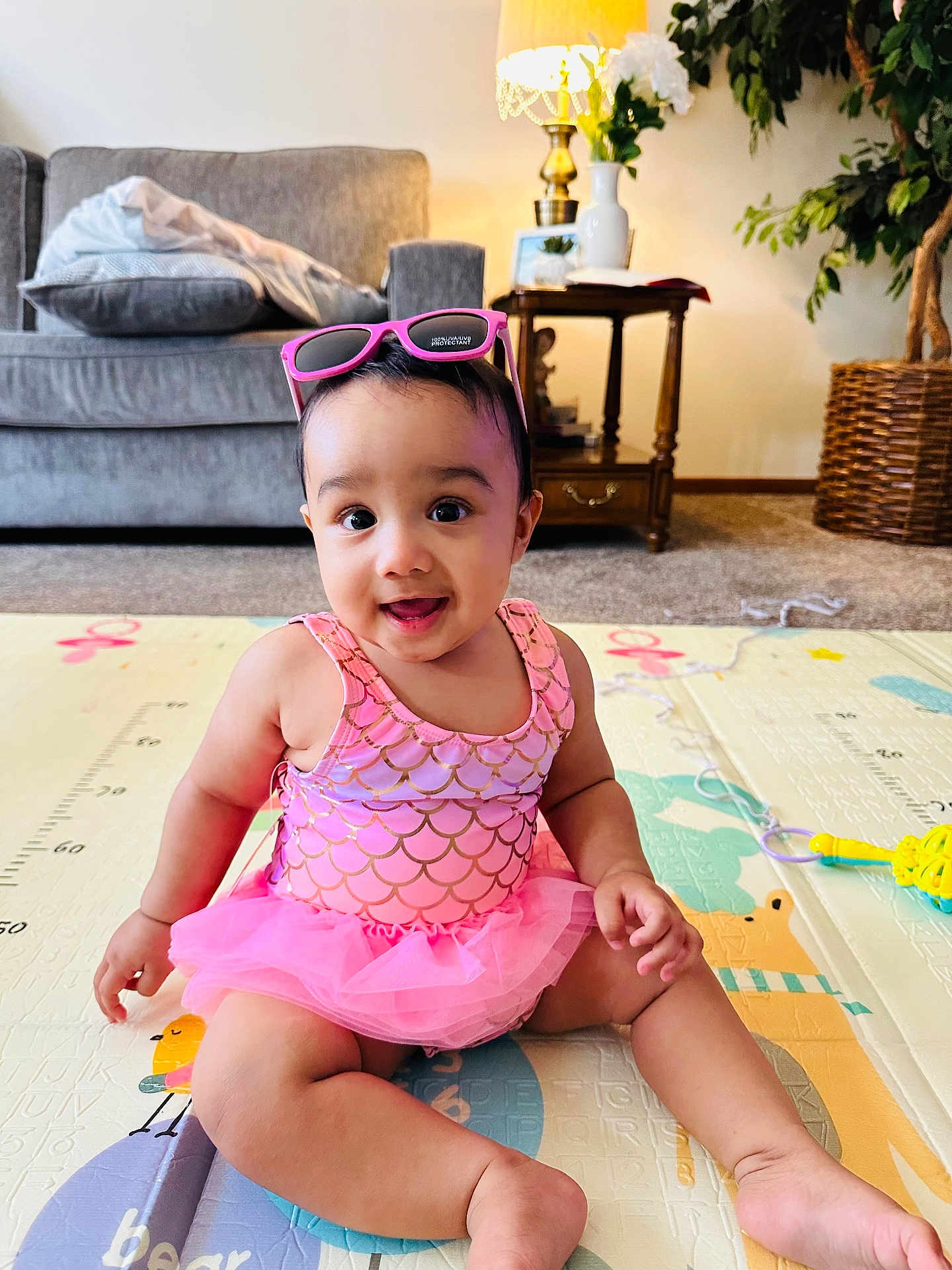Azlin Mehrish is registered to the contest to win money with this photo: baby, child, pink_clothing, sunglasses, play_mat, indoor, smiling, sitting, living_room, lamp, sofa, plant, table, toy, cute, tutu, floor, portrait, happy, young_child
