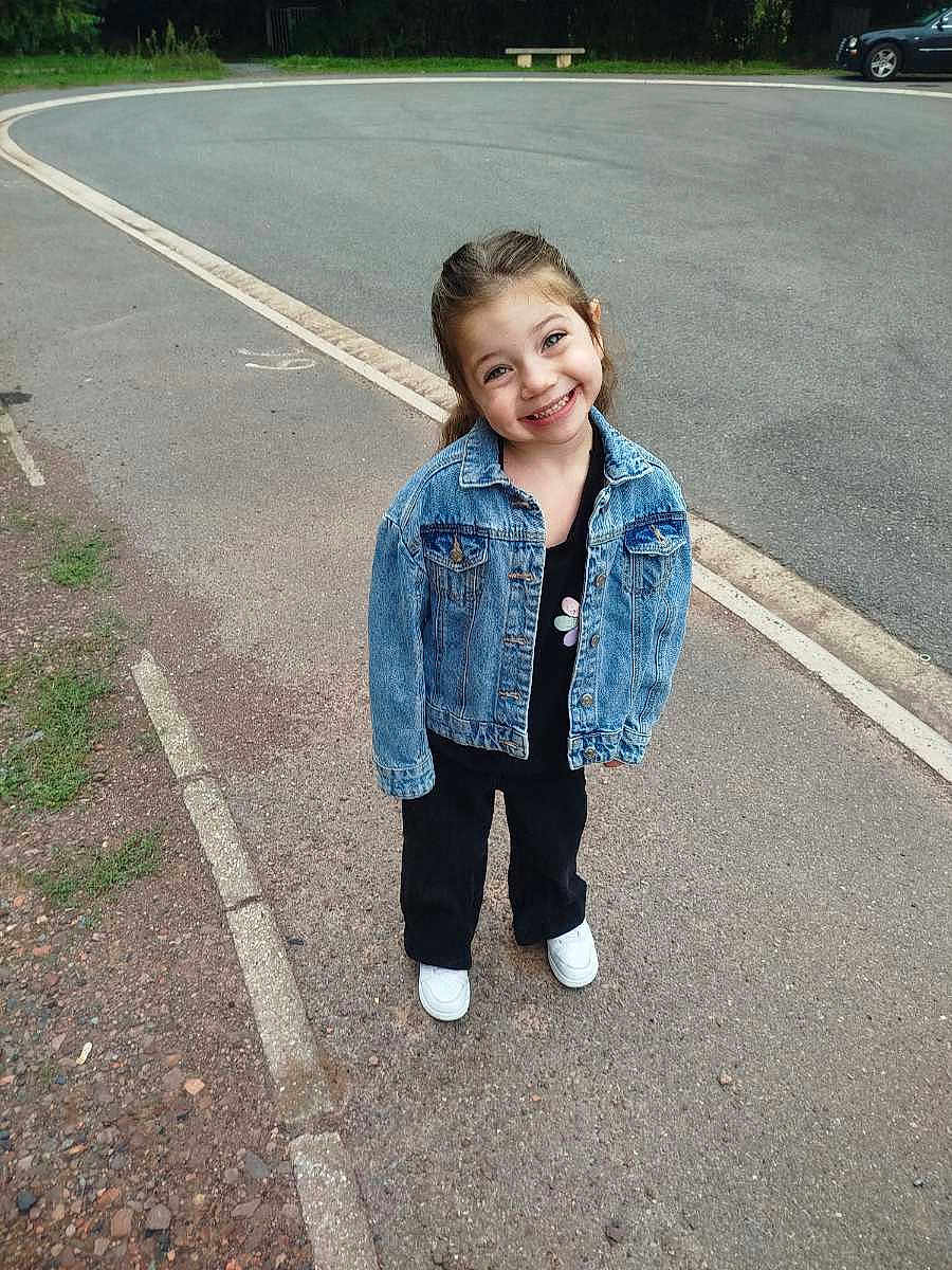 Juditte a rejoint le concours — aidez-le/la à gagner de superbes lots ! black_pants, car, casual_clothing, child, curly_hair, daylight, denim_jacket, girl, happy, nature, outdoor, pavement, person, posing, road, sidewalk, smiling, street, urban, white_shoes