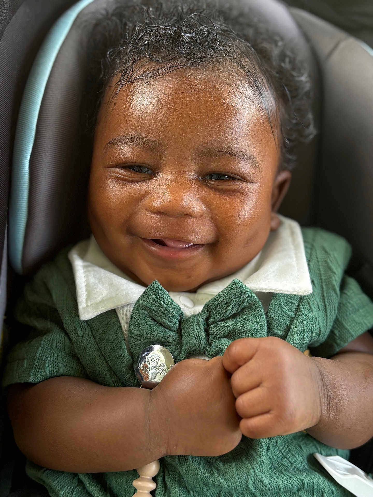 Harlem is registered to the contest to win money with this photo: baby, smile, face, hands, curly_hair, green_clothing, white_collar, car_seat, infant, cute, happy, portrait, closeup, skin, child, adorable, baby_accessory, knitwear, holding, sitting