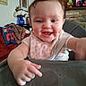 Bella is registered to the contest to win money with this photo: baby, child, high_chair, bib, smiling, dimples, indoor, person, face, hand, table, furniture, home, decor, flower_vase, picture_frame, blanket, clothing, happy, cute
